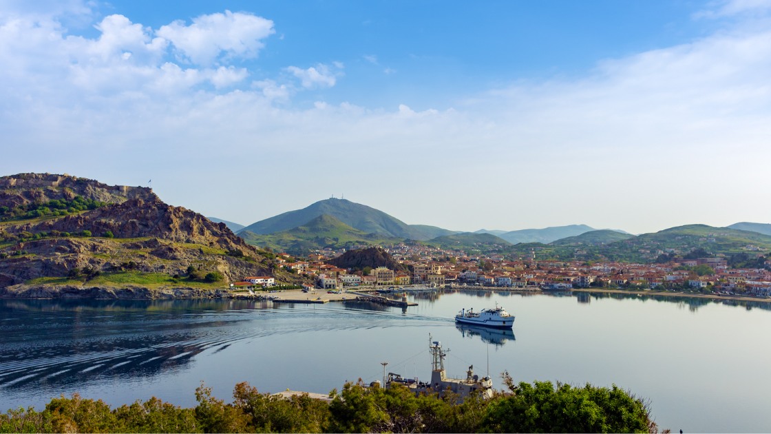 Scenic panorama of Myrina with its harbor, Venetian castle, and surrounding hills, as a ferry arrives at the port.