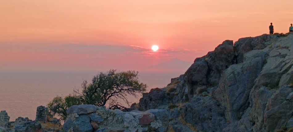 Sunset from Myrina Castle, with rocky cliffs and silhouettes of people against a glowing sky.