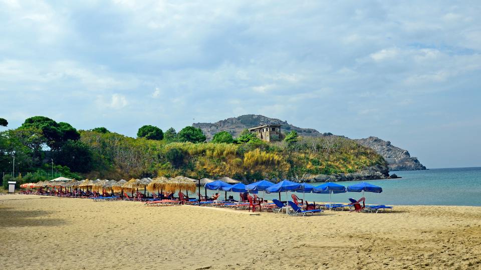 Riha Nera Beach in Lemnos, featuring sandy shores with sunbeds and umbrellas, surrounded by lush greenery.