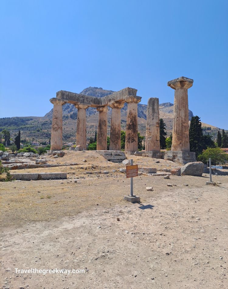 Temple of Apollo ruins in Ancient Corinth with tall columns set against the backdrop of Acrocorinth mountain under a bright blue sky.