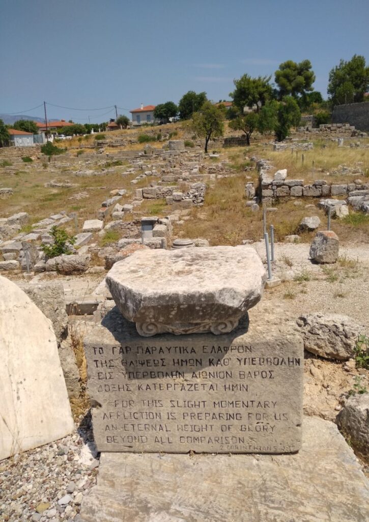 Ancient inscribed stone with a quote from 2 Corinthians, set in a Corinth archaeological site.