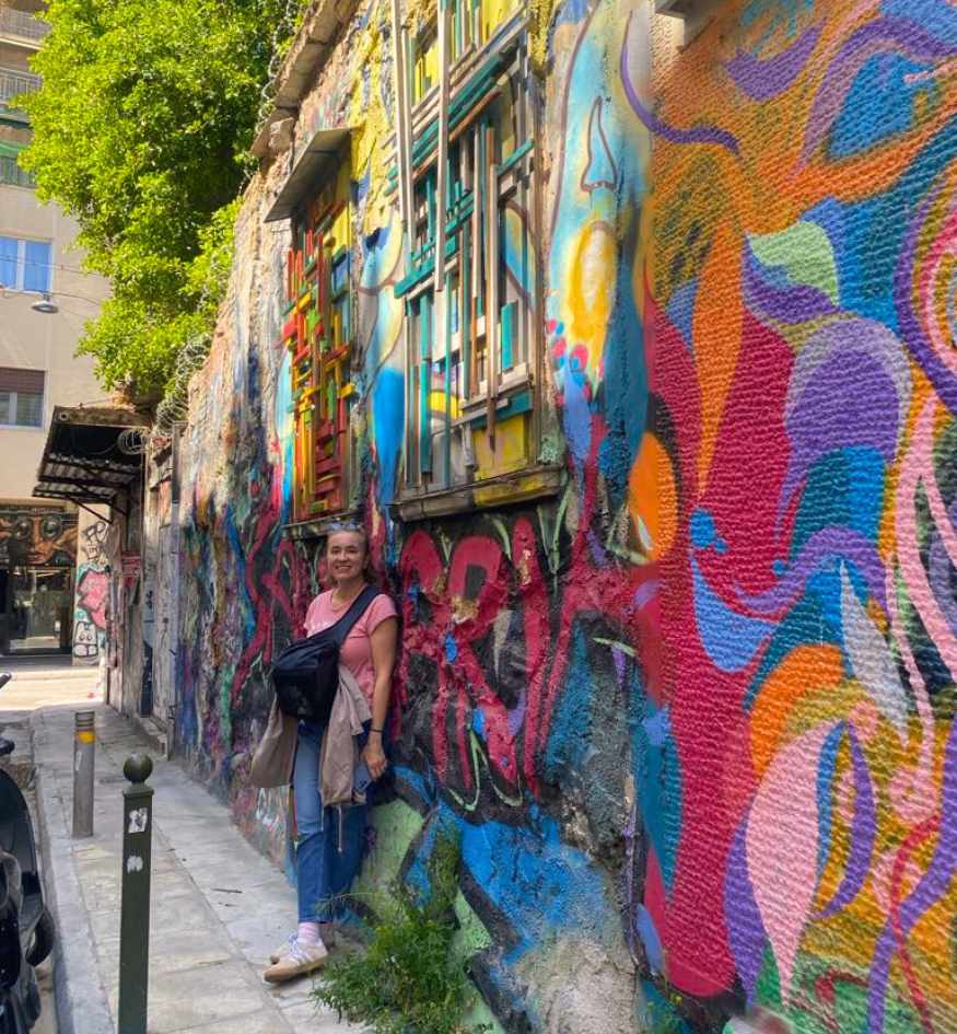 Evgenia in a pink shirt and jeans stands smiling next to a vibrant, multi-colored graffiti mural on a narrow street wall in the Exarchia neighborhood of Athens, Greece.