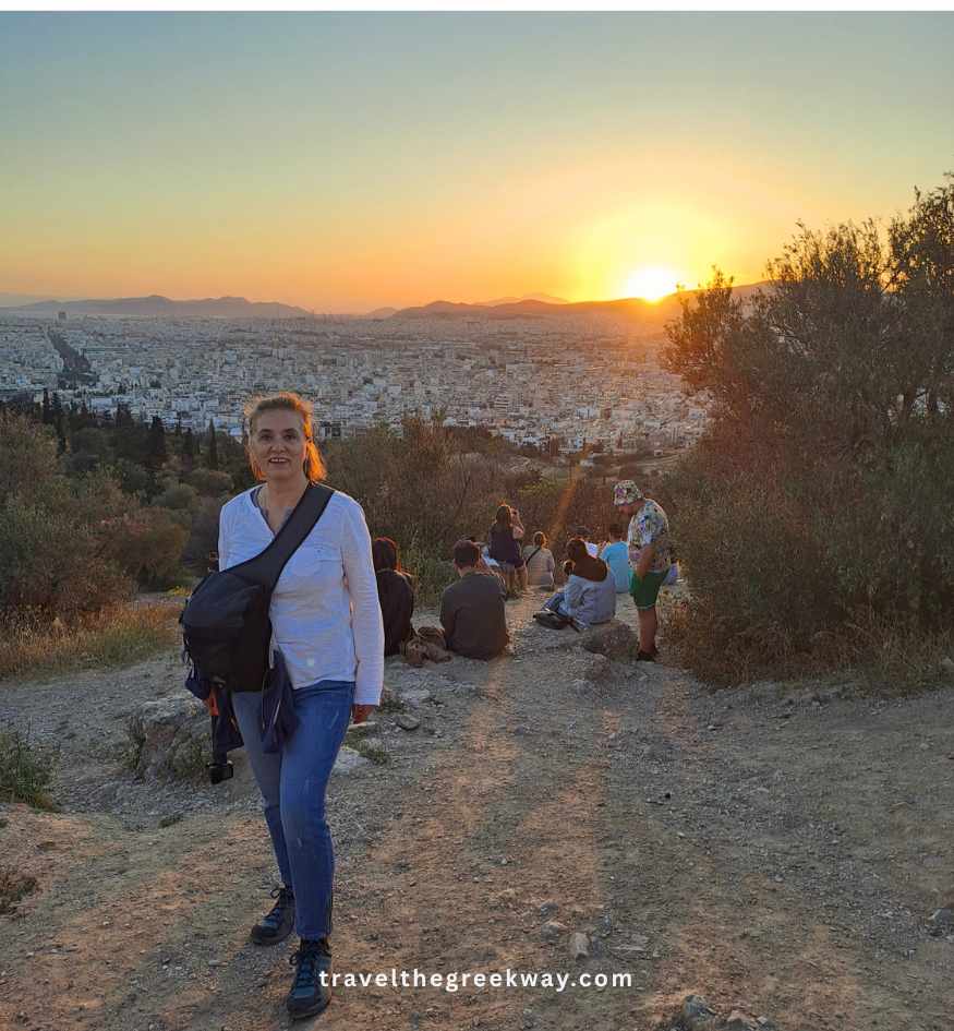 Evgenia stands on a dirt path on Philopappou Hill in Athens, Greece, during a vibrant golden sunset with a crowd of people watching the sun drop behind the distant mountains.