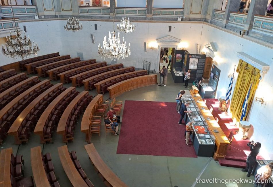 View of the old parliament chamber with wooden benches and chandeliers inside the National Historical Museum of Athens.