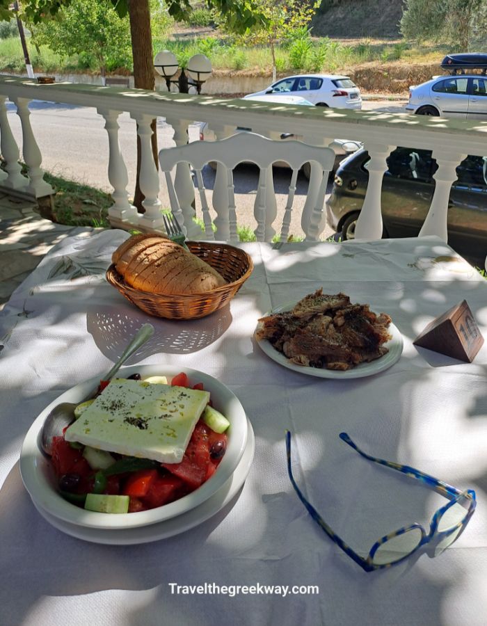Outdoor table with Greek salad, bread, grilled meat, and glasses in a shady taverna in Ancient Olympia Peloponnese.