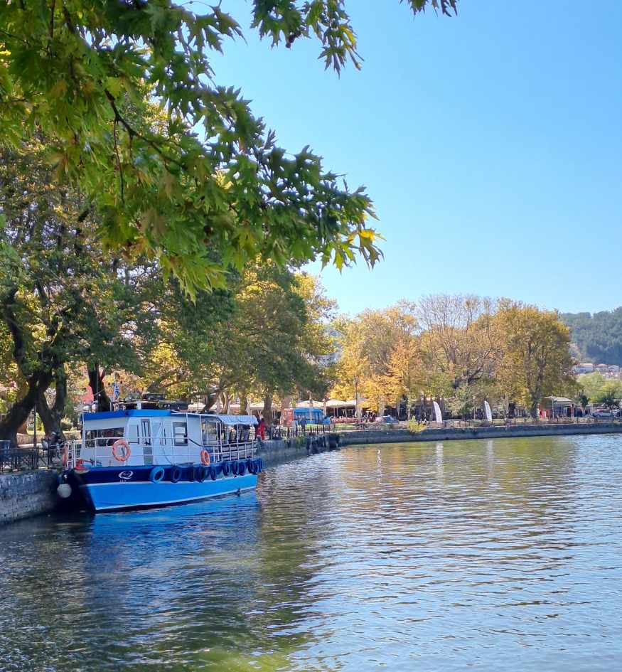 Peaceful waterfront along Lake Pamvotida in Ioannina, Epirus, Greece, with a blue and white passenger boat moored beside a tree-lined promenade, calm lake waters, and outdoor cafés beneath leafy trees.