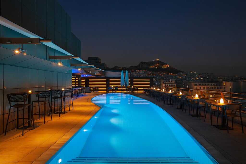 Rooftop pool at night in Athens with Acropolis and city lights in the distance.