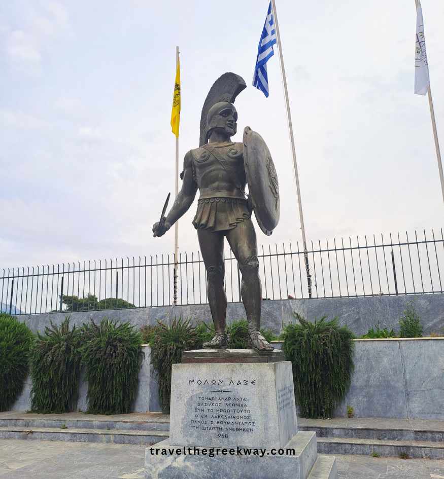 The iconic bronze statue of King Leonidas in Sparta, Peloponnese, standing before the Greek national flag and a blue sky.