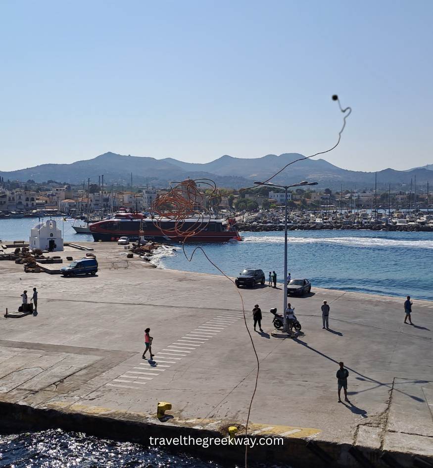 View from a ferry deck of Aegina port, showing a red high-speed boat, a small white chapel, and mountains in the distance.