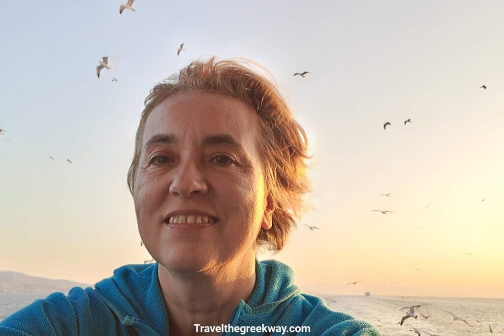 Evgenia of Travel the Greek Way smiling on a ferry deck at sunset with a flock of seagulls flying in the golden sky behind her. Flying vs. ferry to the Greek islands: which is actually better?