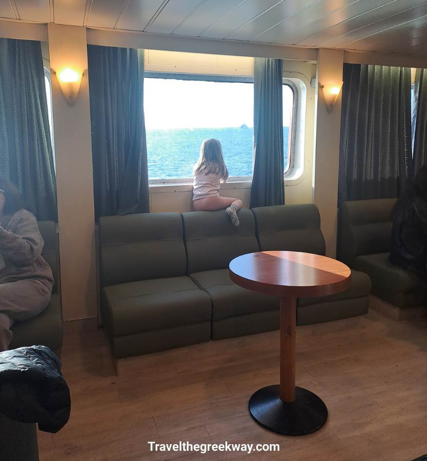 A young child sits on a green sofa inside a ferry lounge, looking out a large window at the blue Aegean Sea.