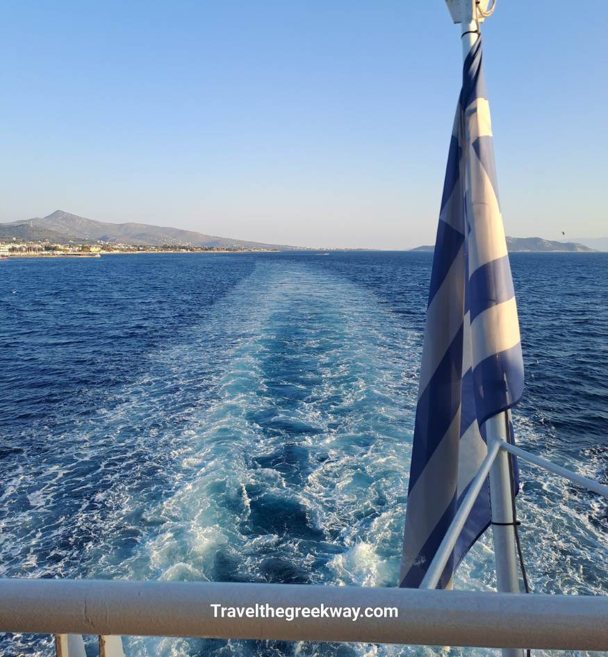 View from a ferry leaving Athens with a Greek flag in the foreground and the sea wake trailing behind.