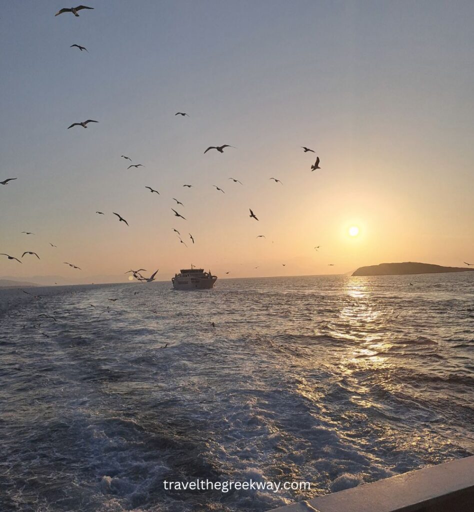 Ferry at sunset in Greece with seagulls flying over the sea, golden light on the water, and an island silhouette in the distance.