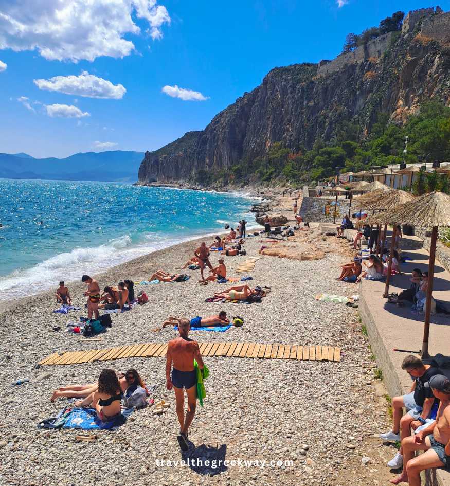 People sunbathing on a pebbly beach in Nafplio, Greece, with a towering cliff and the blue Argolic Gulf in the background.