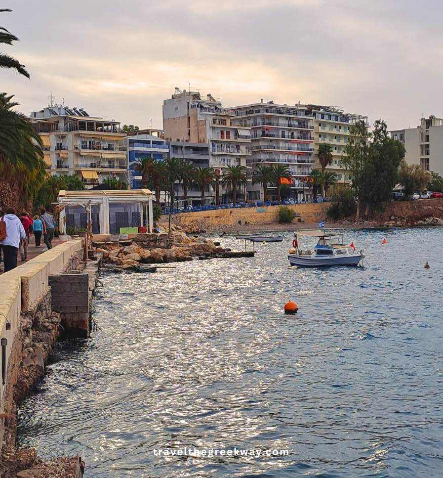 Small boats moored in the blue waters along the Loutraki waterfront promenade with seaside buildings in Peloponnese, Greece.
