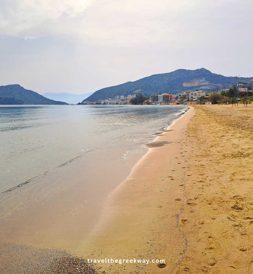 A long, quiet stretch of golden sand at Tolo beach, with calm blue water and coastal hills under a soft, hazy sky.