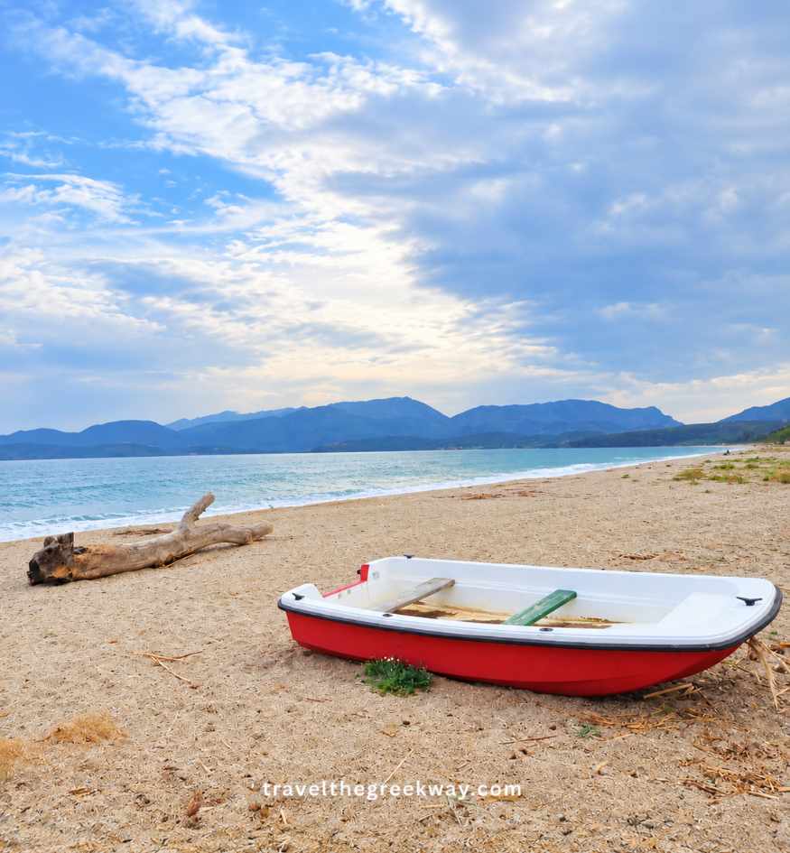 Small red-and-white fishing boat resting on Mavrovouni Beach near Gythio, with the sea and mountains of the Mani Peninsula in the background.