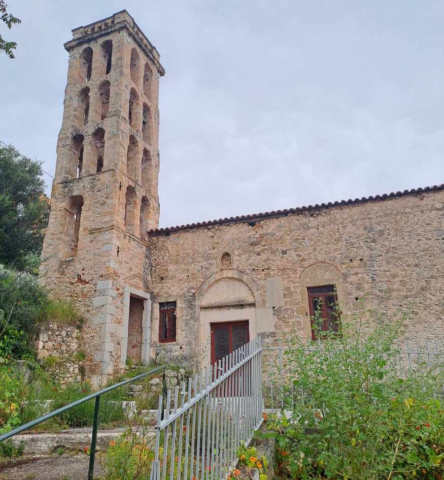 Historic stone church with a tall weathered bell tower in Gythio, Greece, surrounded by greenery.