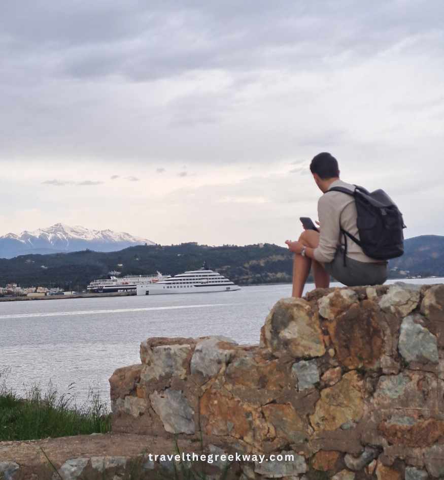 Visitor sitting on the stone walls of Kranae Island overlooking the sea, Gythio harbor and distant mountains.