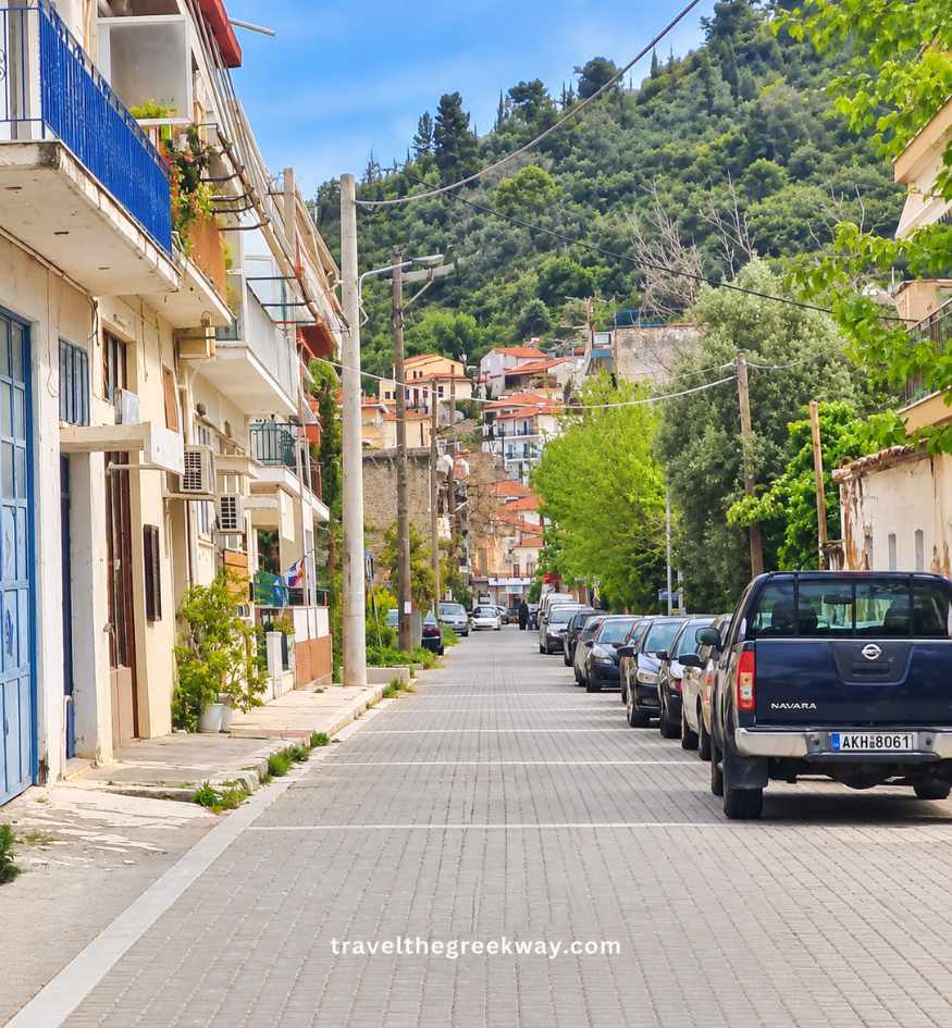 Quiet residential street in Gythio with traditional hillside houses, parked cars and a green wooded hill rising behind the town.