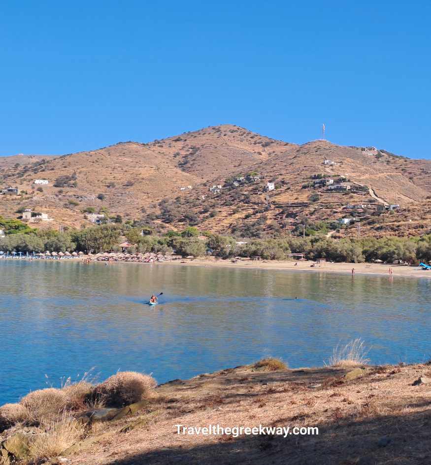 Wide view across the calm blue bay of Otzias Beach, with a sandy shore and dry terraced hills rising behind it on Kea island, Greece.