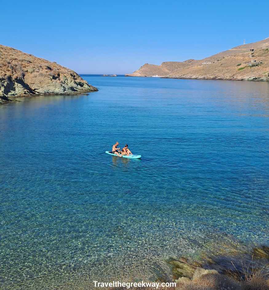 Crystal-clear turquoise water at Otzias Beach on Kea island, Greece, with two people on a paddleboard and rocky hills surrounding the bay.