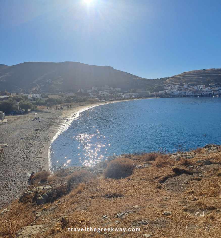 View across Korissia Beach and its curved bay, with calm blue water and the port village of Korissia in the distance on Kea, Greece.