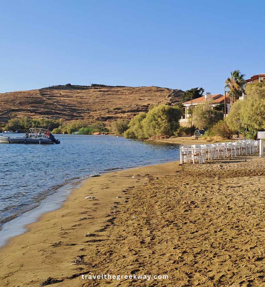 Sandy waterfront in Vourkari with outdoor tables, small boats and low hills surrounding the peaceful bay on Kea island, Greece.