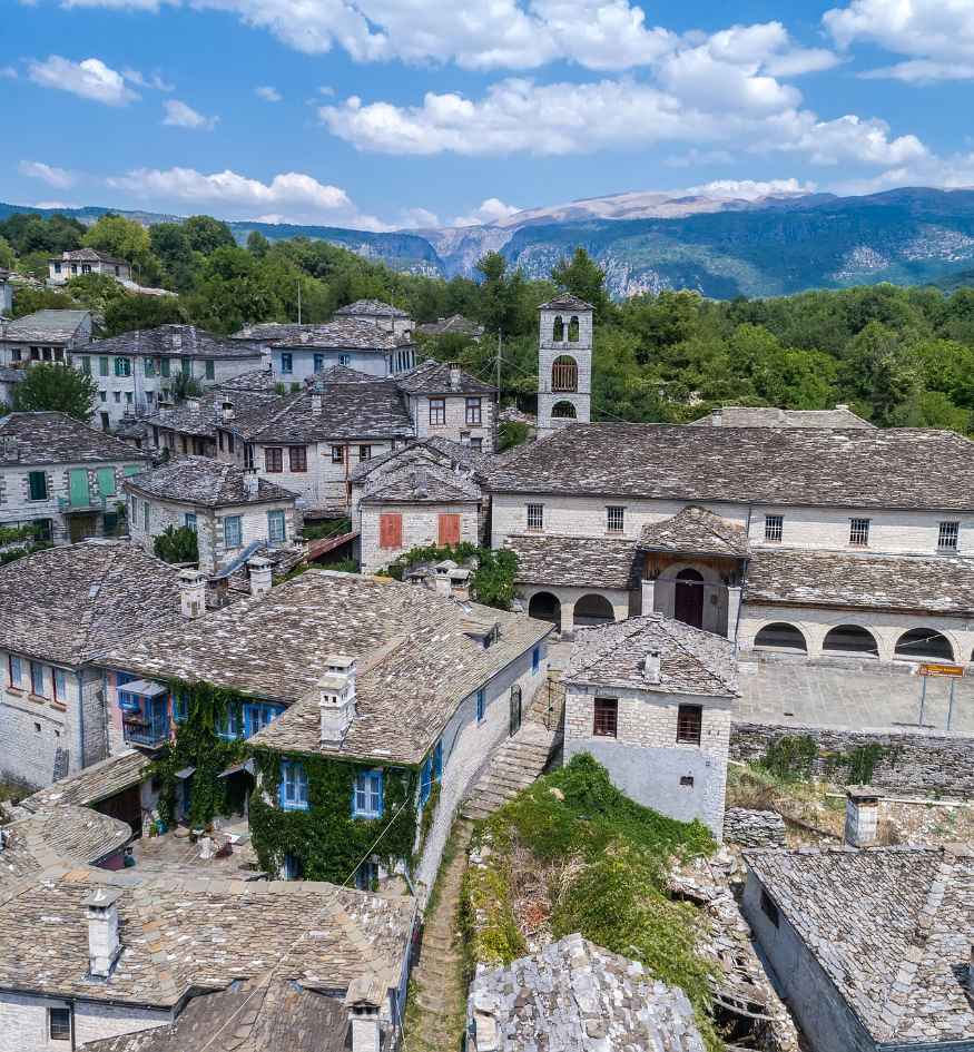 Panoramic view of Dilofo village in Zagori, Epirus, Greece, showing traditional stone houses with gray slate roofs, a historic church with a bell tower, and forested mountains rising behind the village under a partly cloudy sky.