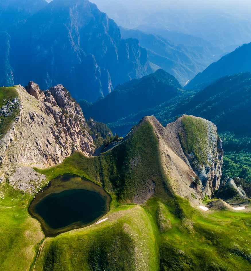 Aerial view of Drakolimni Dragon Lake in the mountains of Zagori, Epirus, Greece, surrounded by steep grassy slopes, rocky peaks, and dramatic mountain ranges stretching into the distance.