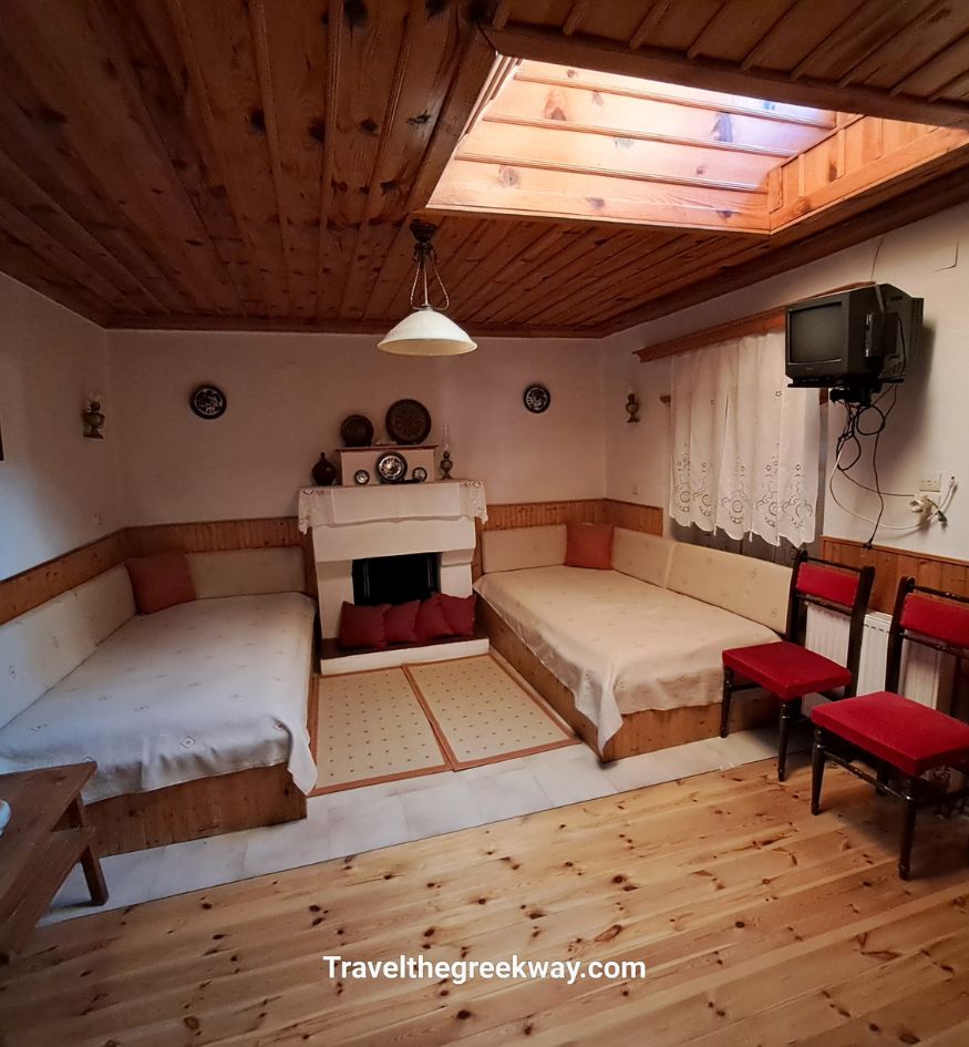 Cozy interior of a traditional guesthouse in Tsepelovo, Zagori, Greece, featuring wooden floors and ceilings, two built-in sofa beds, a small fireplace, red chairs, and a skylight providing natural light.