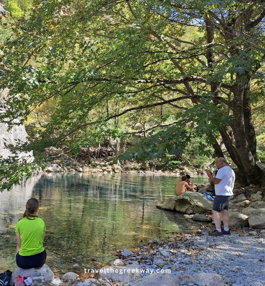 Visitors relaxing beside the crystal-clear waters of the Voidomatis River in Vikos Gorge, Zagori, Greece, surrounded by large rocks, leafy trees, and a shaded pebble-covered riverbank.