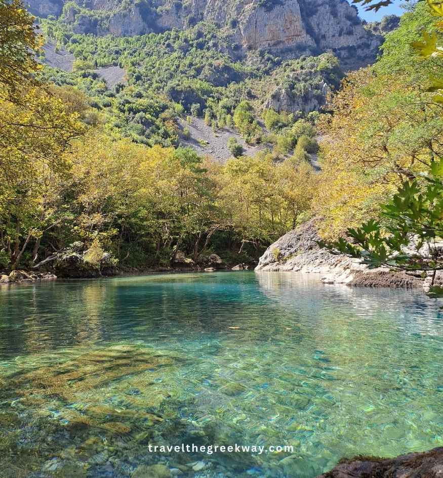 Stunning turquoise waters of the Voidomatis River in Zagori, Epirus, Greece, with a rocky riverbed visible through the clear water, surrounded by forested slopes, autumn-colored trees, and towering limestone cliffs.