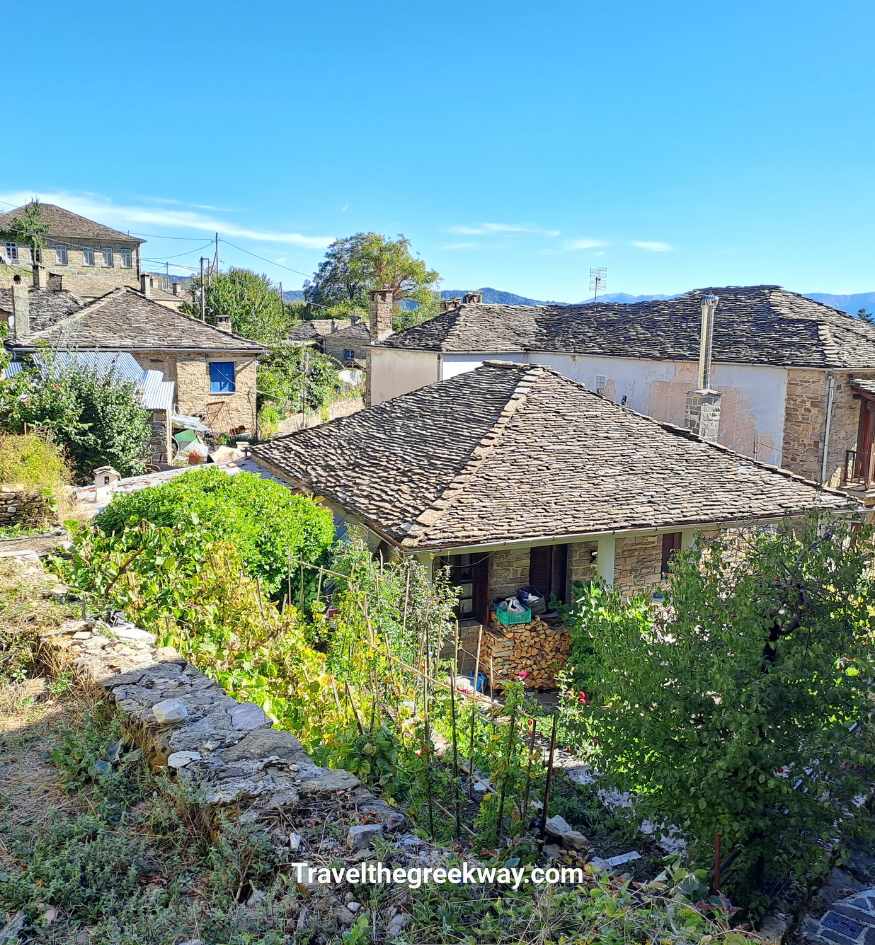 Traditional stone houses in Tsepelovo village, Zagori, Greece, featuring characteristic gray slate roofs, stone chimneys, small gardens, and green vegetation, with mountain peaks visible in the distance beneath a clear blue sky.